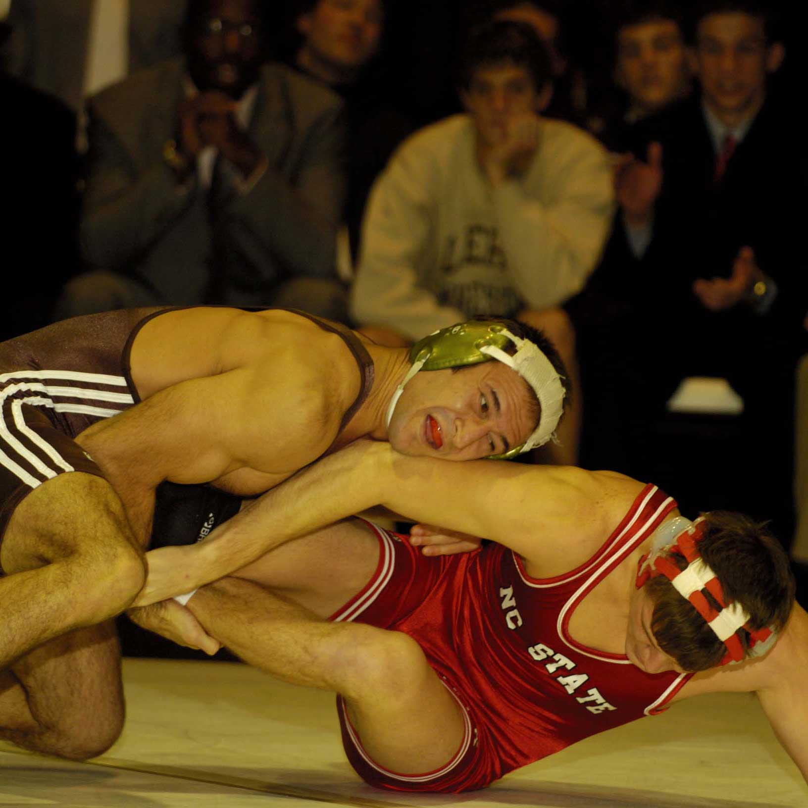 Travis wears his wrestling uniform and red mouthguard. He wrestles his NC State opponent to the mat while pulling their legs.