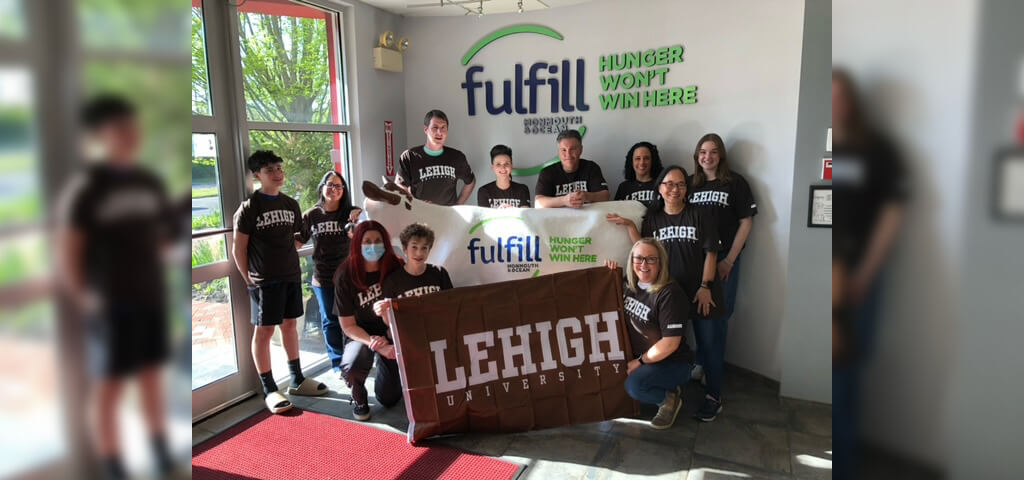 Holding a Lehigh banner and a "Fulfill" banner, a group wearing brown Lehigh tshirts pose in front of the "Fulfill" sign