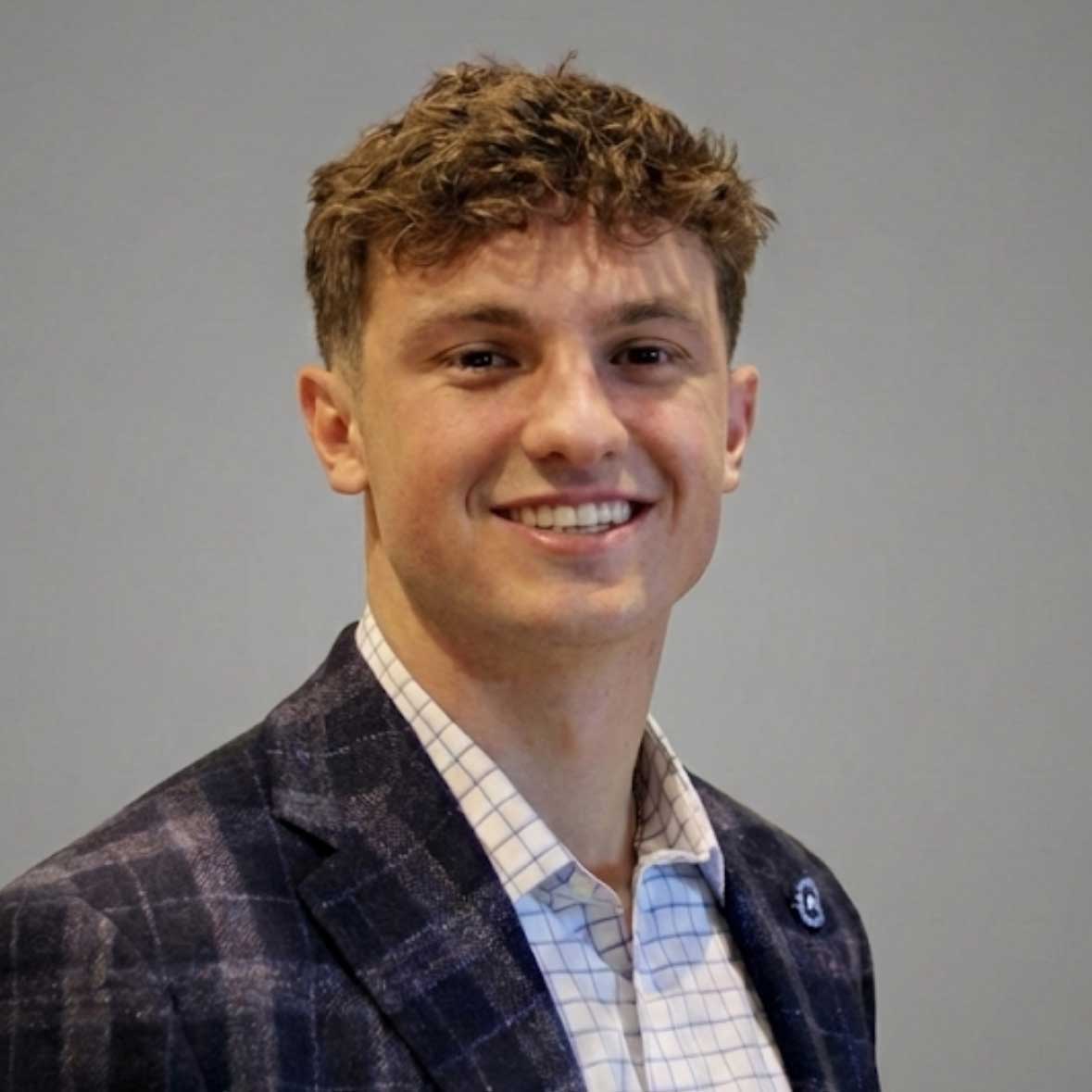 Tyler wears a checkered suit jacket with a white button up underneath. He stands in front of a gray background and smiles.