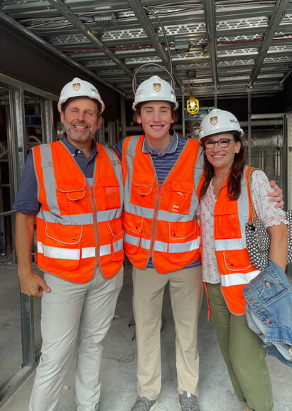 Doug, George, and Elissa Tiesi stand side-by-side, wearing orange construction vests and hardhats inside a construction site.