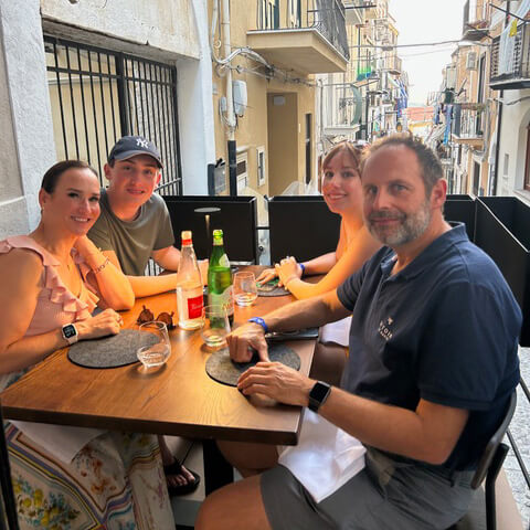Seated at an Italian outdoor bistro, a family of four with mother, father, and adult son and daughter turning to smile for a photo.