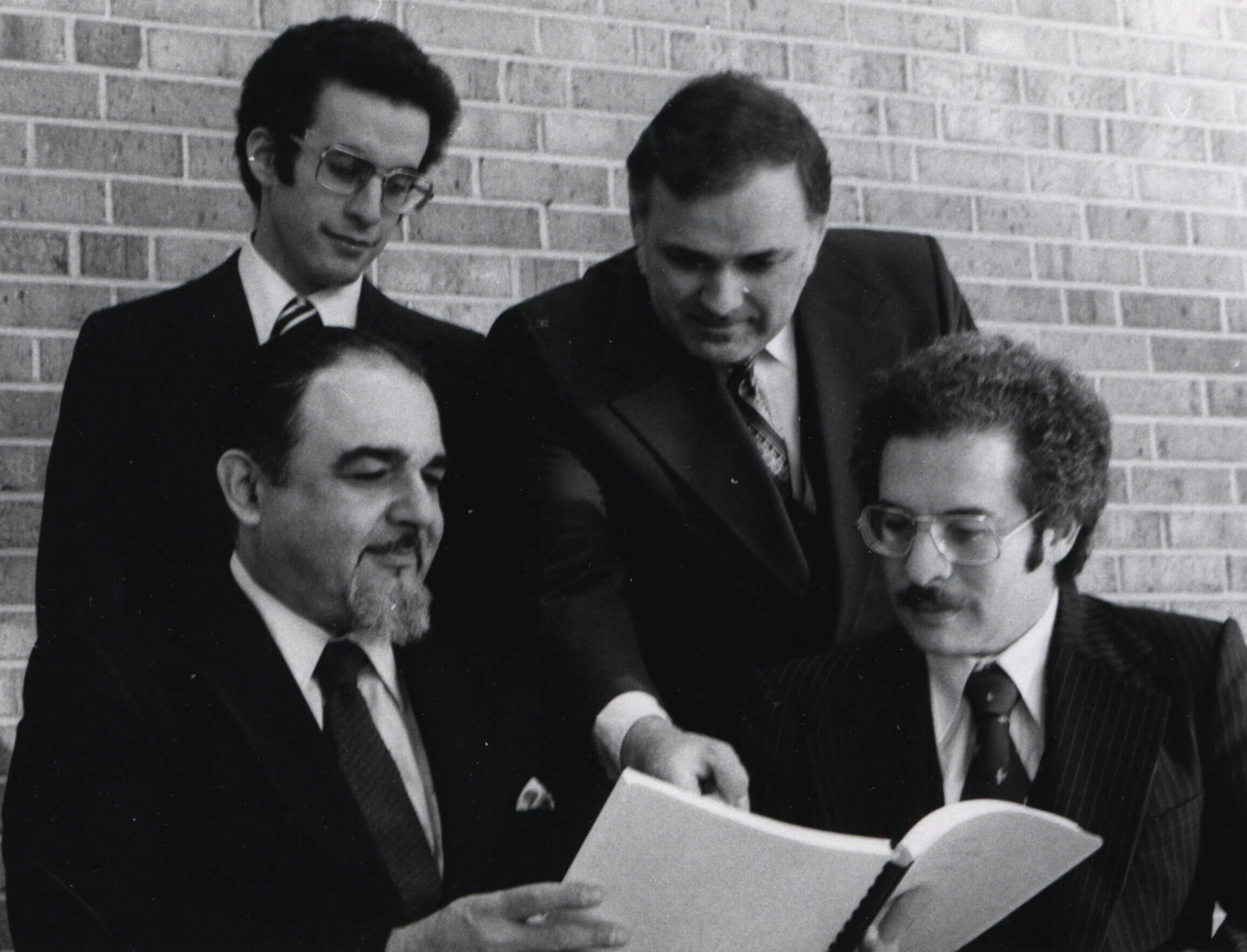 With a brick wall behind them, David Van Horn leans over the shoulder of two men holding a booklet and points to something in the book as another man stands to his left.