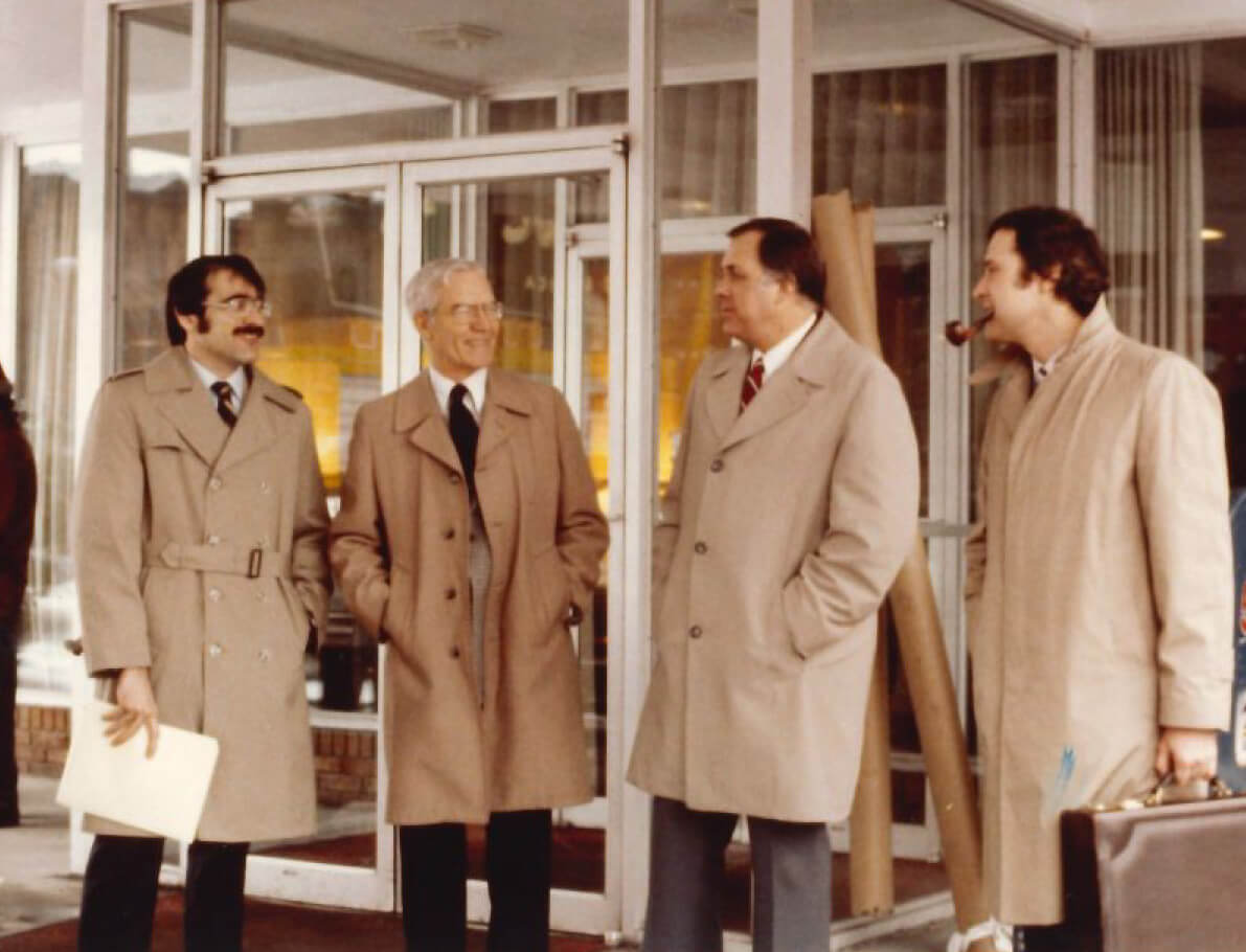 All wearing long tan trench coats, David Van Horn stands with his hands in his pockets outside glass doors to a building with three other male faculty members.
