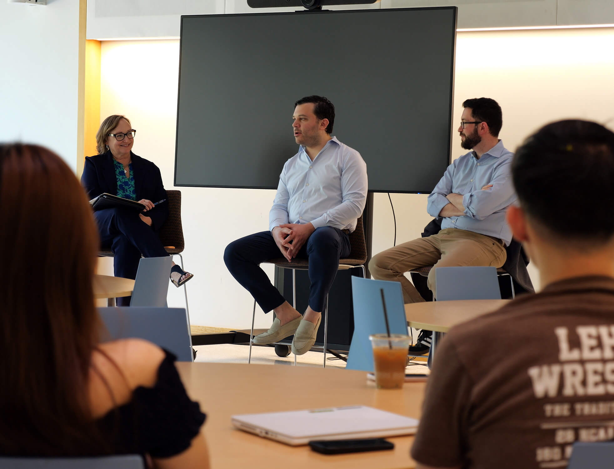 Seen over the shoulders of student attendees, two Vytalize founders sit at the head of a room on stools beside Dean Beth Dolan.