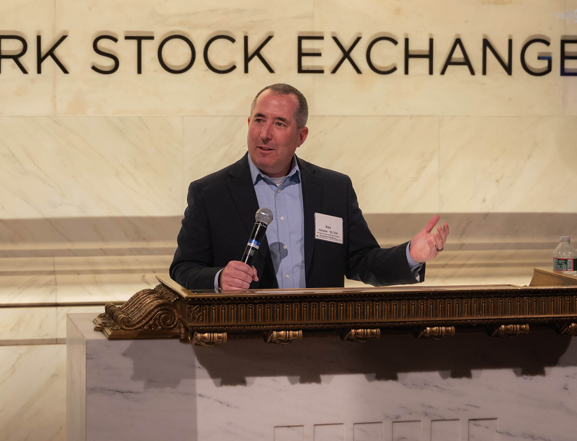 Ken Hoexter speaks at the New York Stock Exchange podium, holding a microphone in one hand and gesturing with the other.