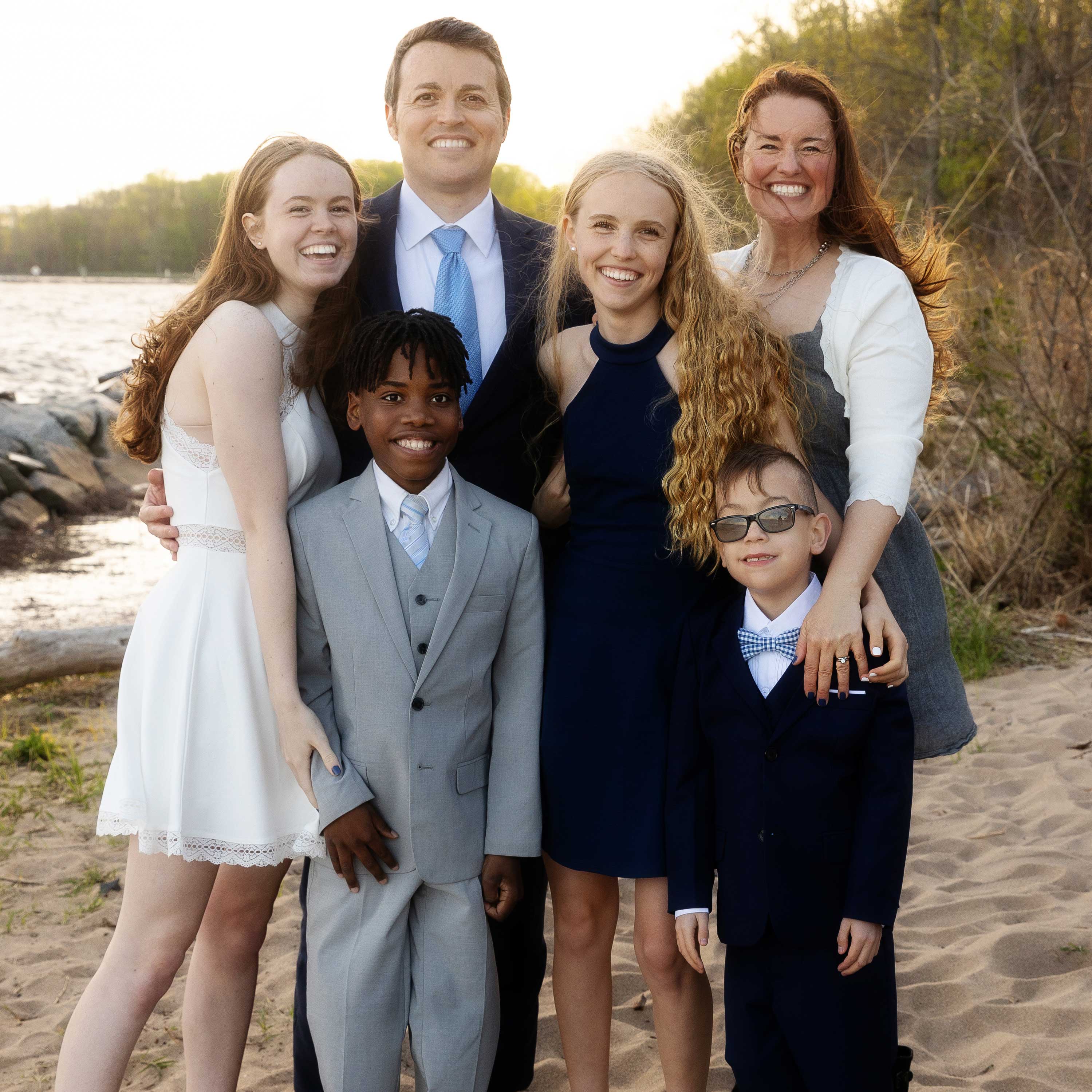 Kelly and Kevin stand with their four children on a beach during golden hour. They all wear business attire and smile warmly.