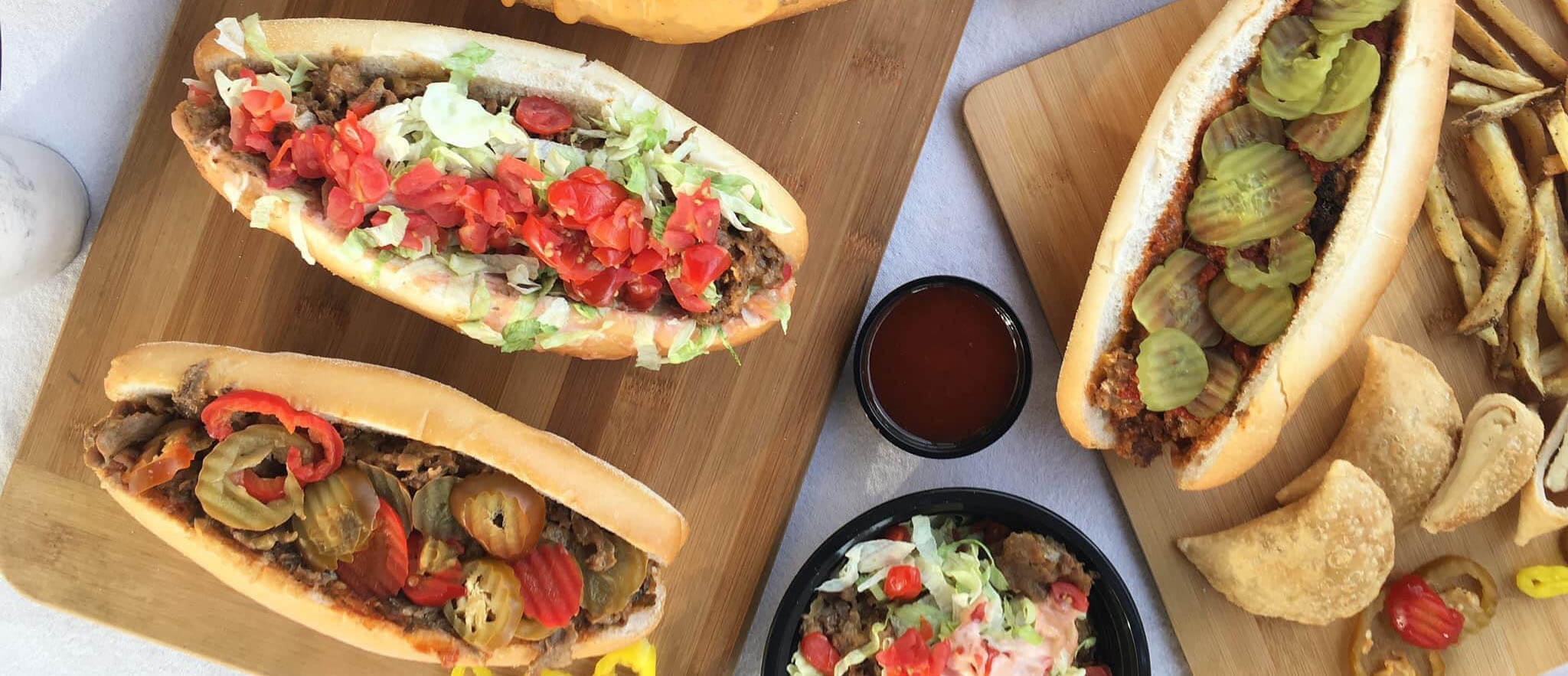 Three cheesesteak sandwiches topped with pickles, lettuce, and tomatoes on wooden cutting boards with french fries on the side
