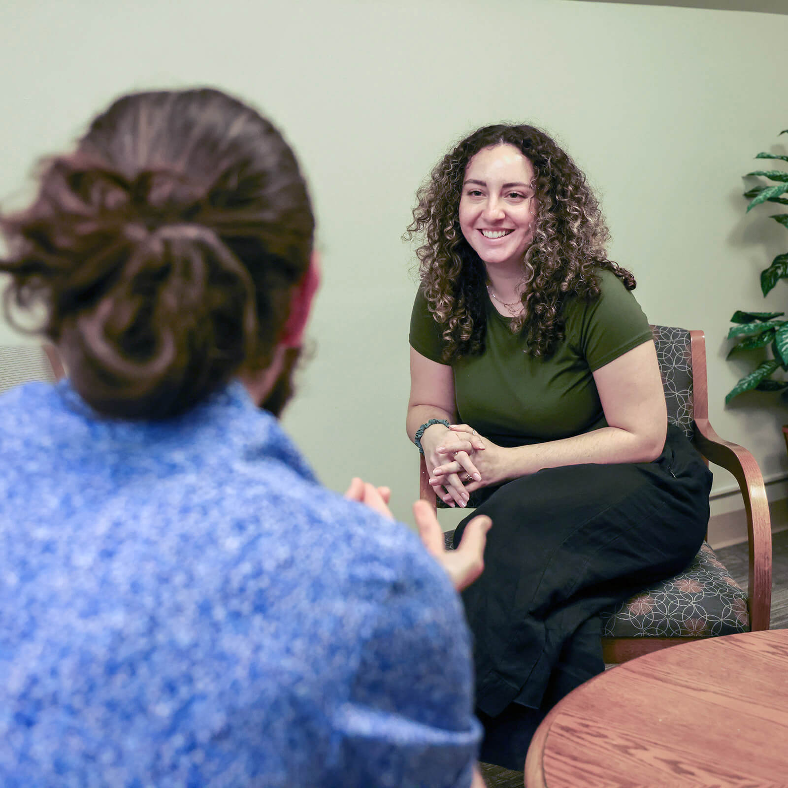 Jessica Stafi, wearing a dark green tshirt and black pants, sits facing a student whose back is facing the camera.