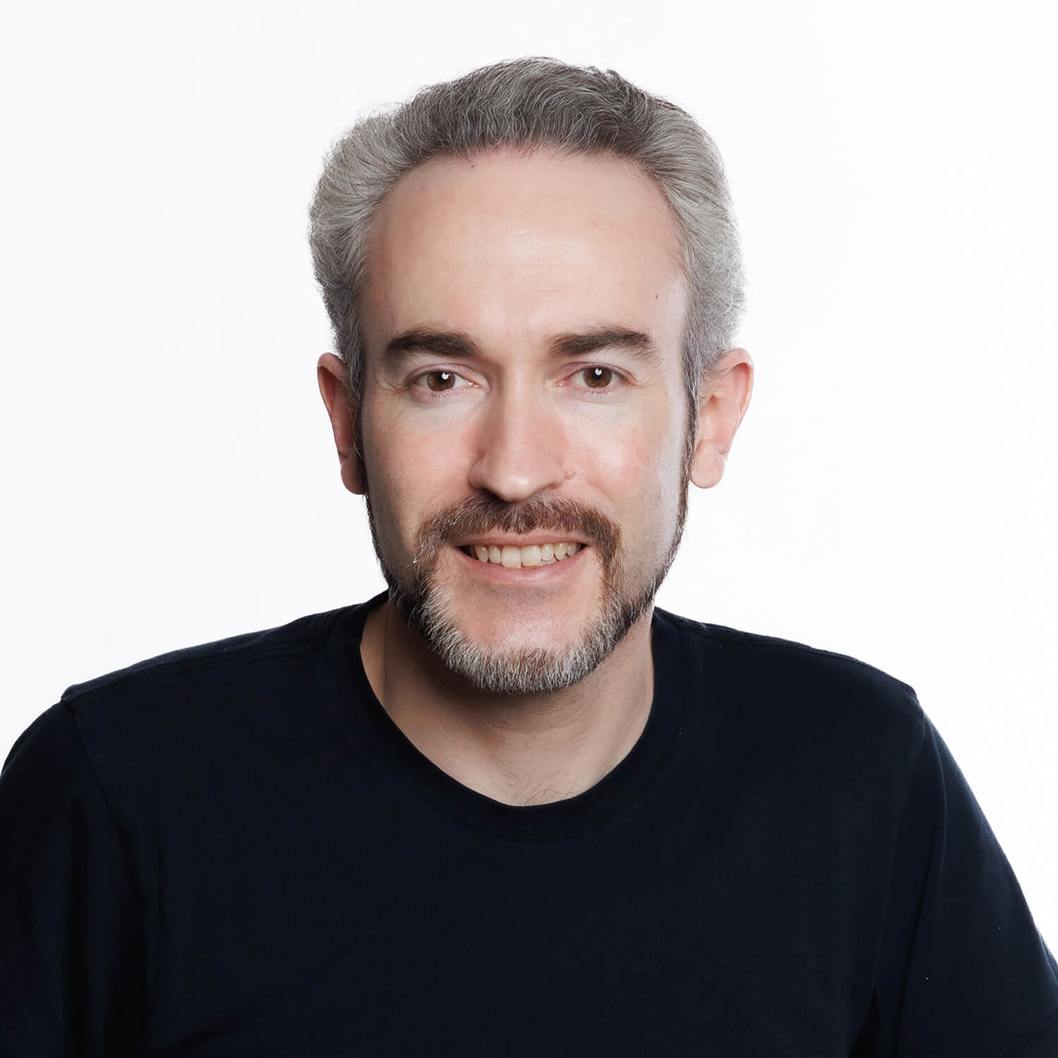 Against a white background, a man smiles for a headshot wearing a black crewneck sweater and a well trimmed beard.