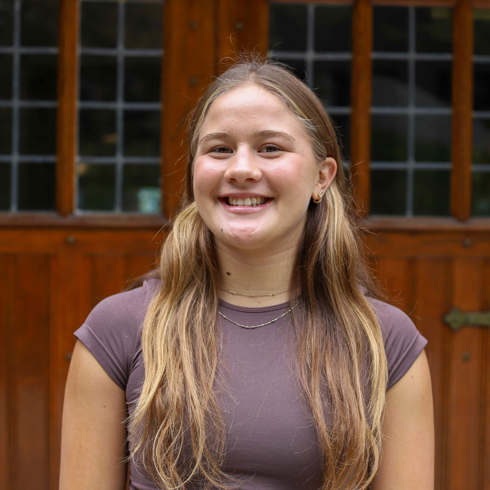 Zorina wears a purple t-shirt and a silver necklace and smiles. Door to a building on campus are visible behind her.