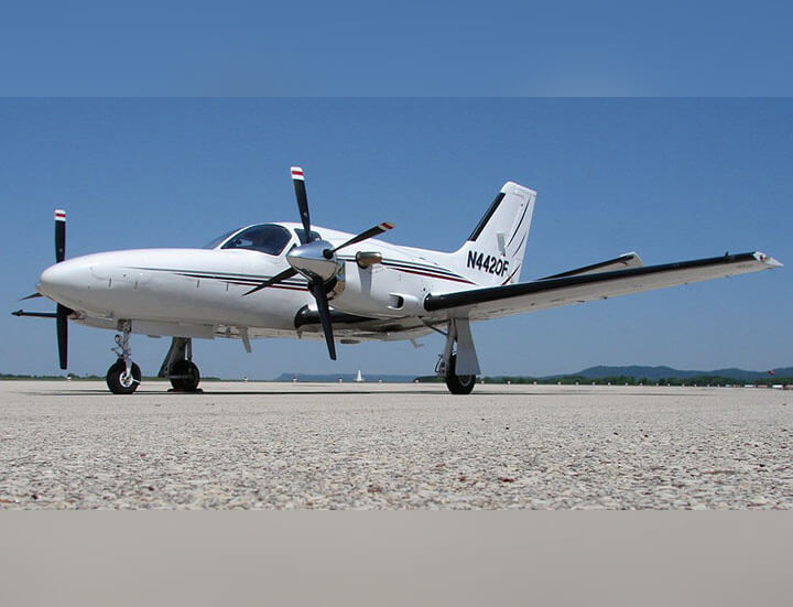 A twin-engine turboprop with 2 propellers and designed to seat five to seven passengers sits parked on the ground with clear skies and low mountains in the distance.