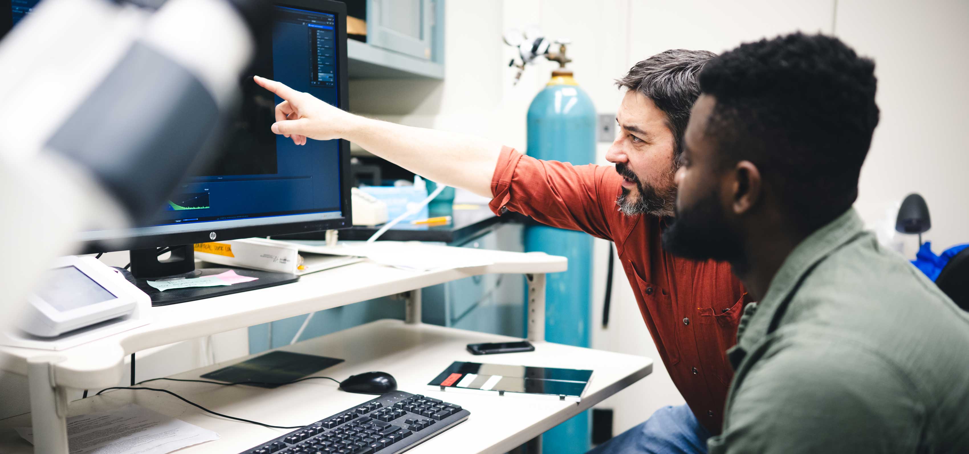 Two men sit at a desk while looking at a computer screen.