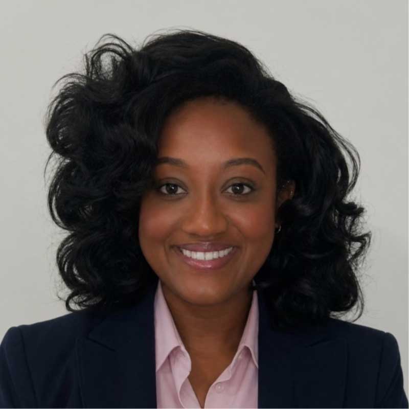 Michaela wears a dark blazer with a pink button up underneath. She stands in front of a gray background and smiles.