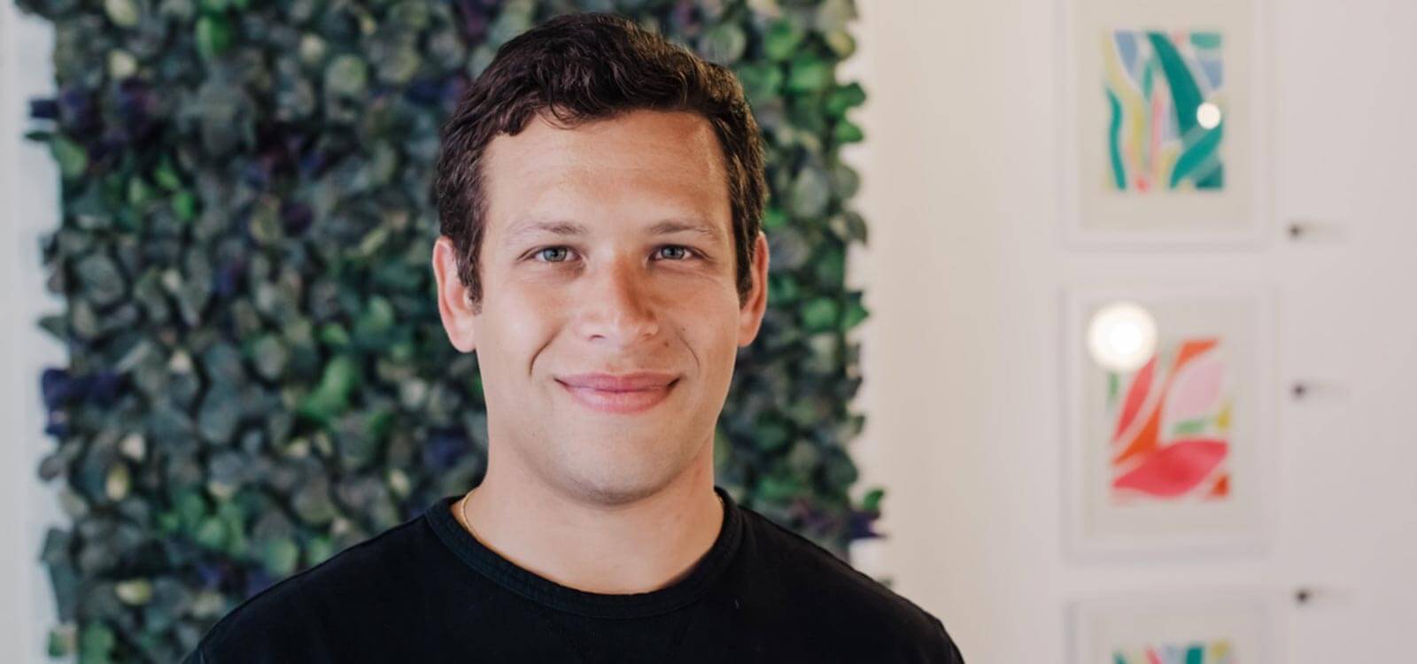 Goldstein wears a black shirt and smiles in a warm, bright room with greenery behind him.