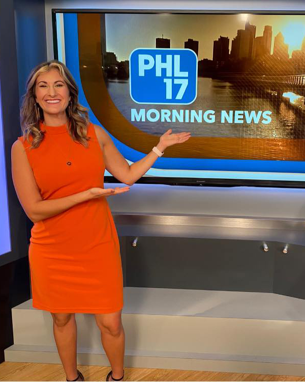 Ariella smiles while wearing a bright orange dress. She gestures to a screen beside her that reads 'PHL 17 Morning News.'