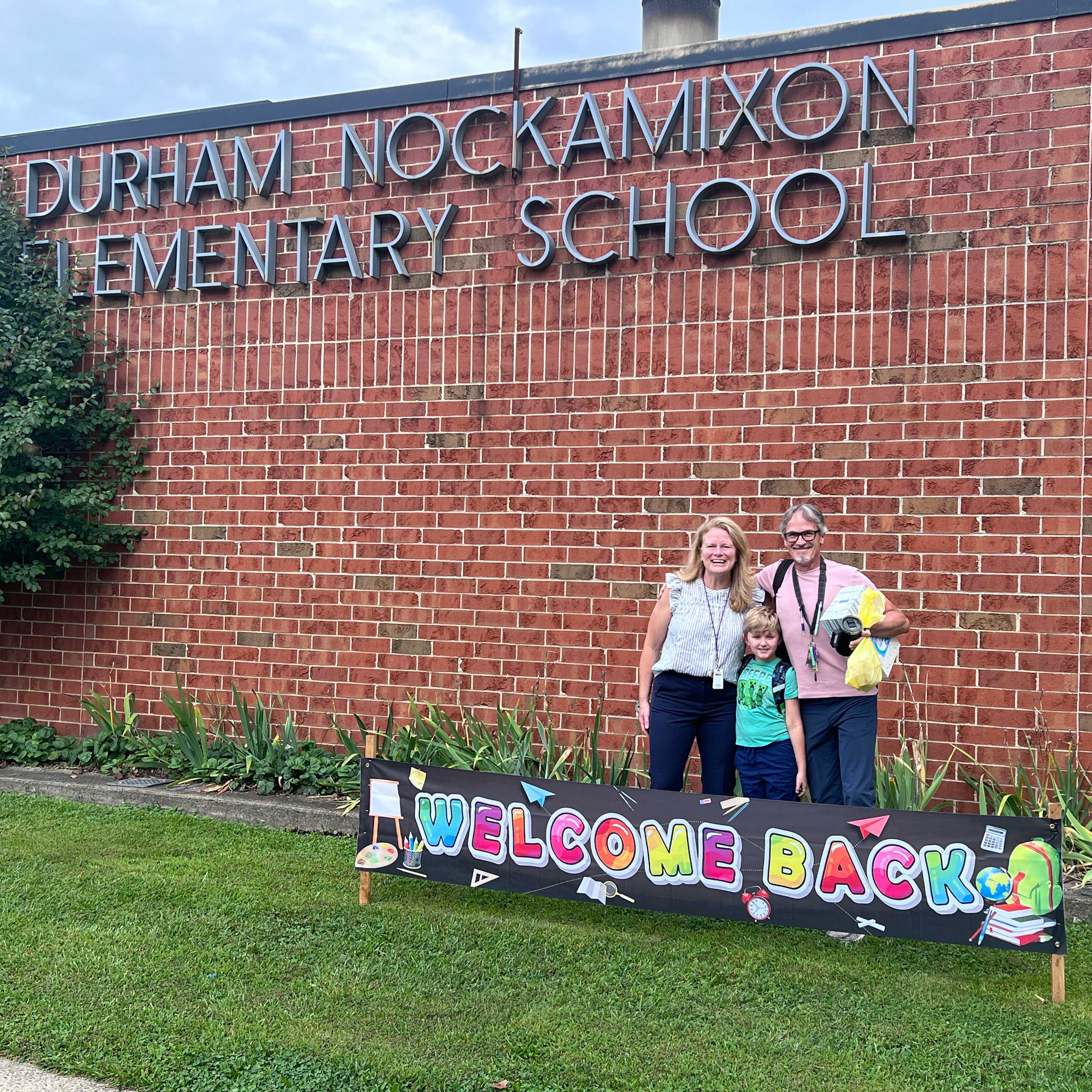 Bridget stands in front of Durham Nockamixon Elementary School next to a 'welcome back' banner; she's with a student and an adult.