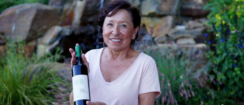 A woman in front of a garden with a rock waterfall holding a bottle of wine while smiling.