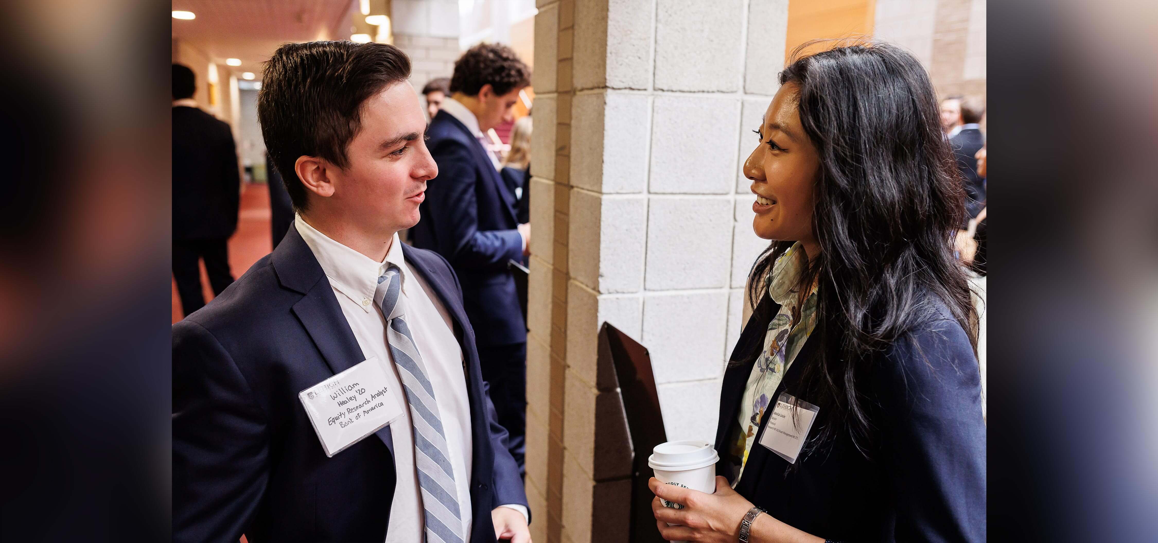 Billy wears business casual and stands facing another Lehigh alumni in similar attire; they both wear name tags.