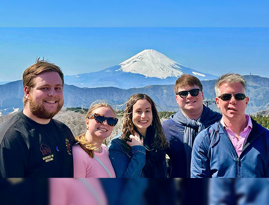 A family of five smile in front of a scenic view of a mountain on a sunny day.