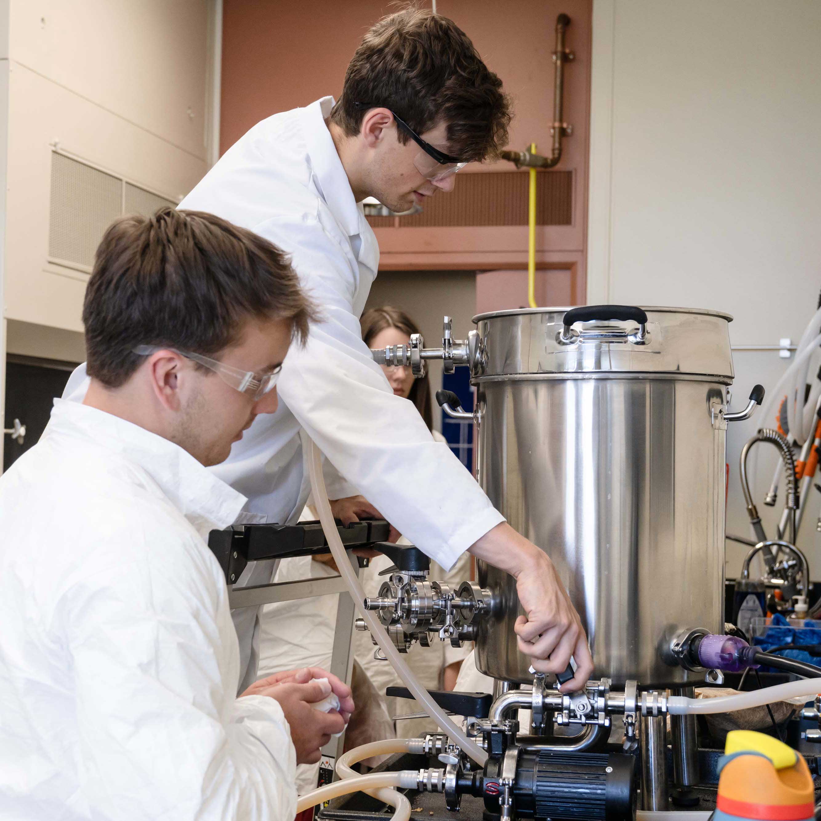 Three students in white lab coats lean over a stainless steel fermentation tank. One student leans forward and adjusts a lever.