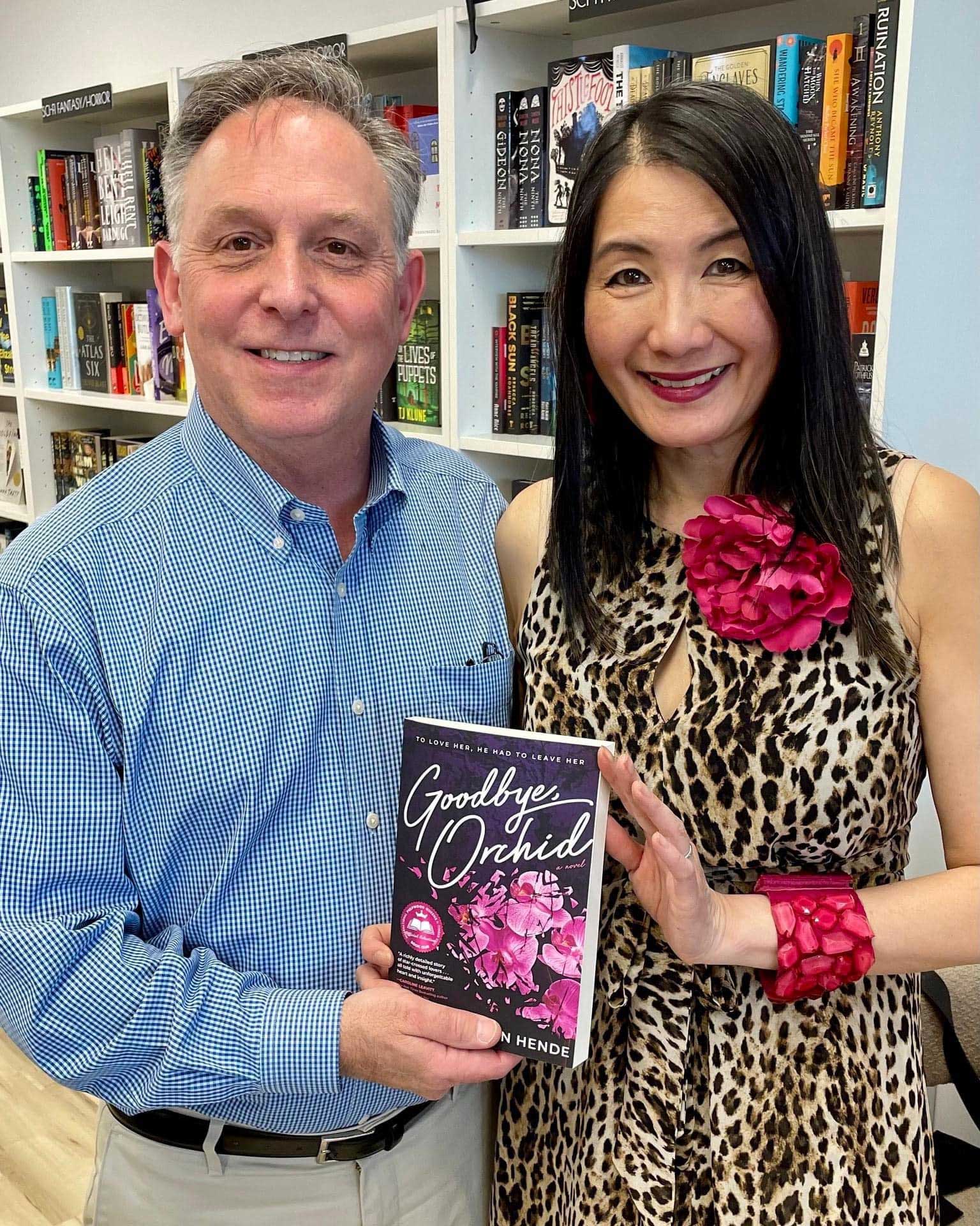 Carol and her husband stand in a bookstore and hold up her novel 'Goodbye, Orchid.' Carol wears a leopard dress and orchid pin.