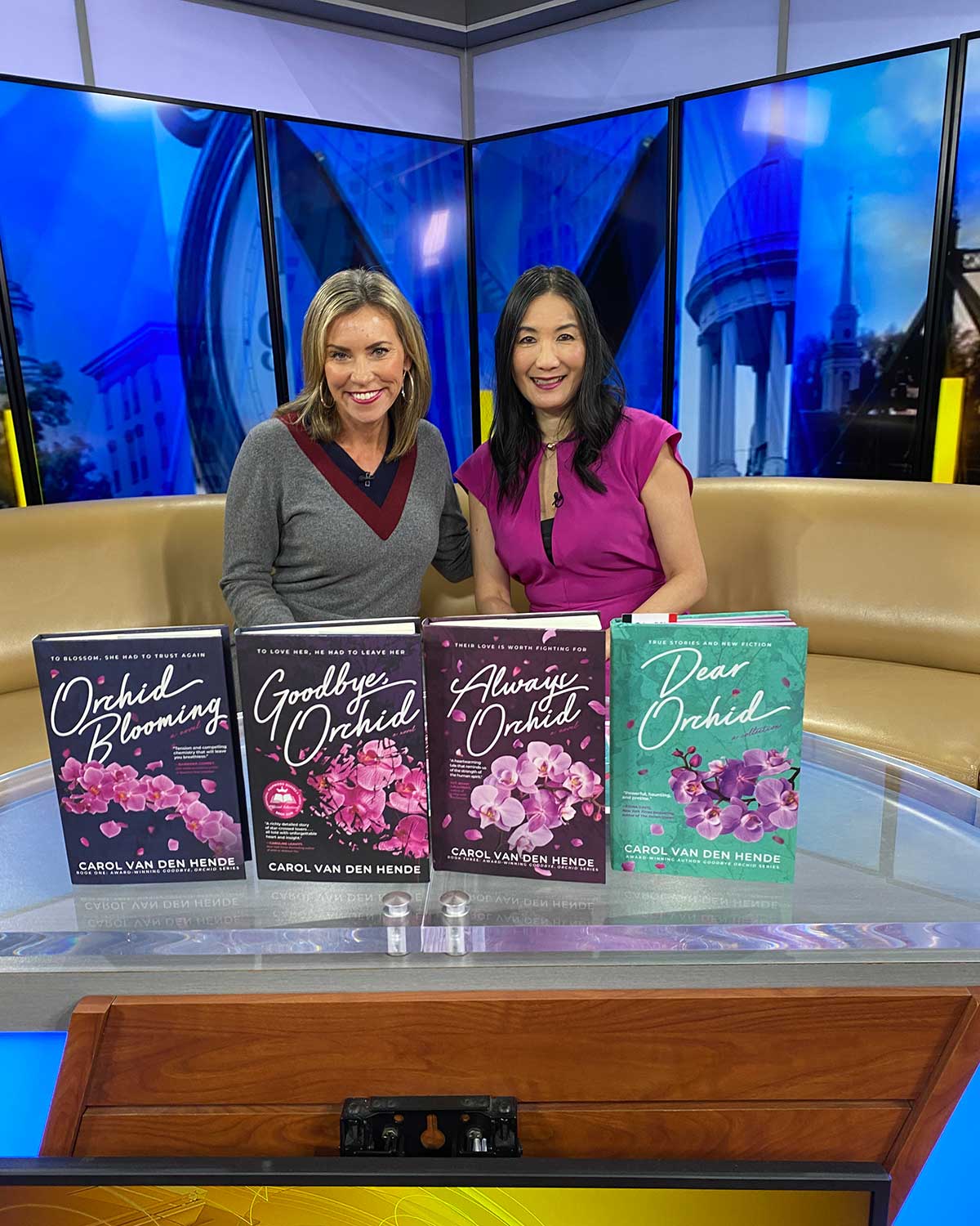 Carol sits in the WFMZ newsroom with an anchor to her left and her novels in the foreground. Carol wears a bright pink blouse.