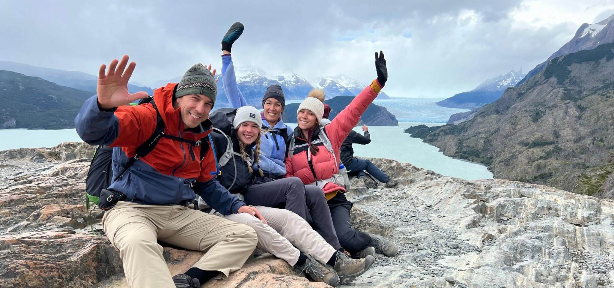 Chris and his family wear brightly colored jackets and sit on top of a mountain. The four of them smile and throw their hands up.