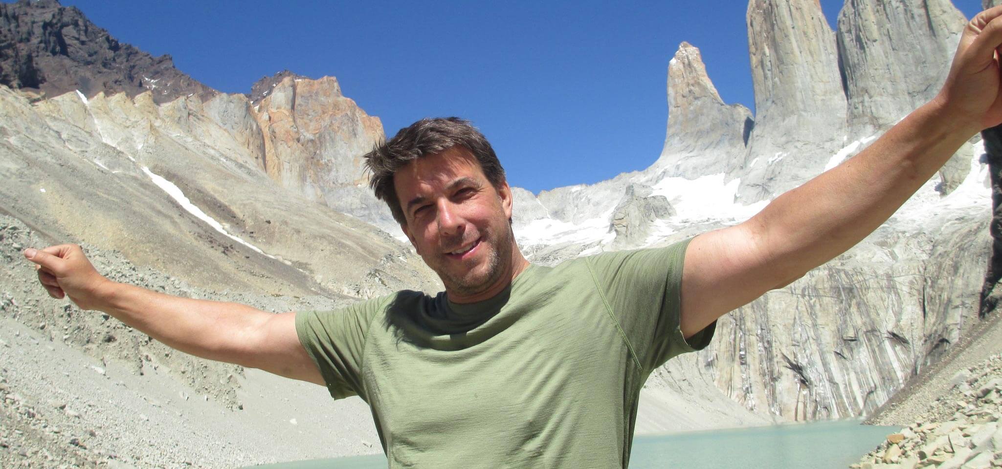 Chris wears a light green t-shirt and stands in the mountains with his arms spread wide.