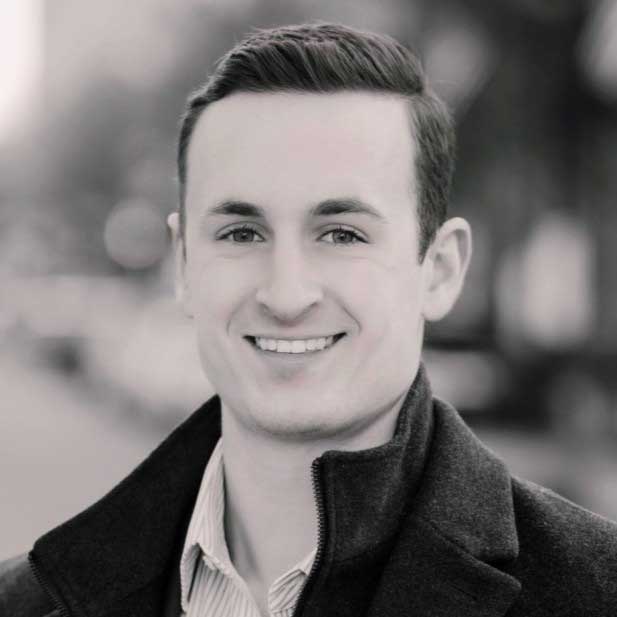 A black and white photo of Chris smiling. He wears a dark zip-up jacket white a striped white button down underneath.