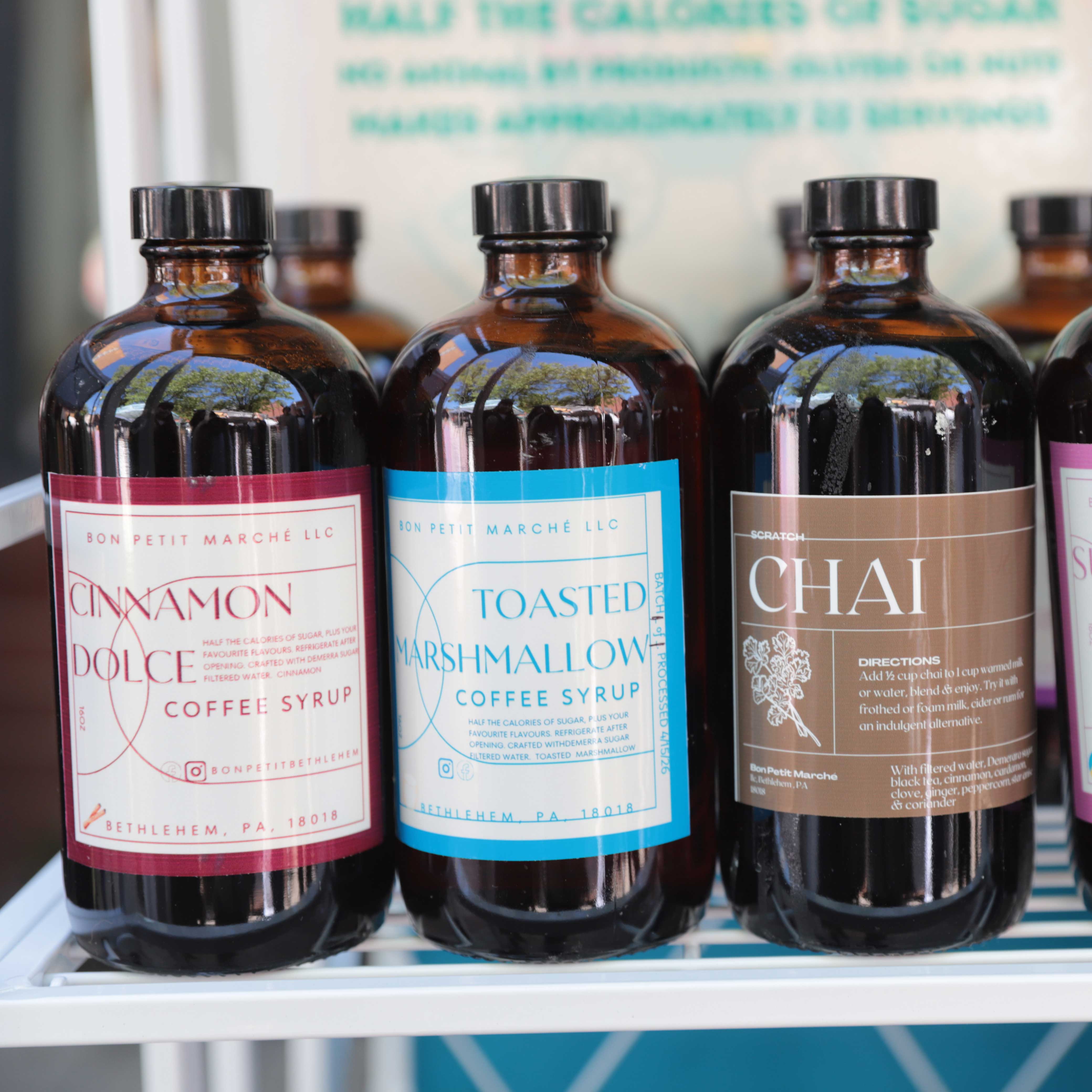 Three dark, glass bottles beside each other on a stand: cinnamon dulce syrup, toasted marshmallow syrup, and chai concentrate. 