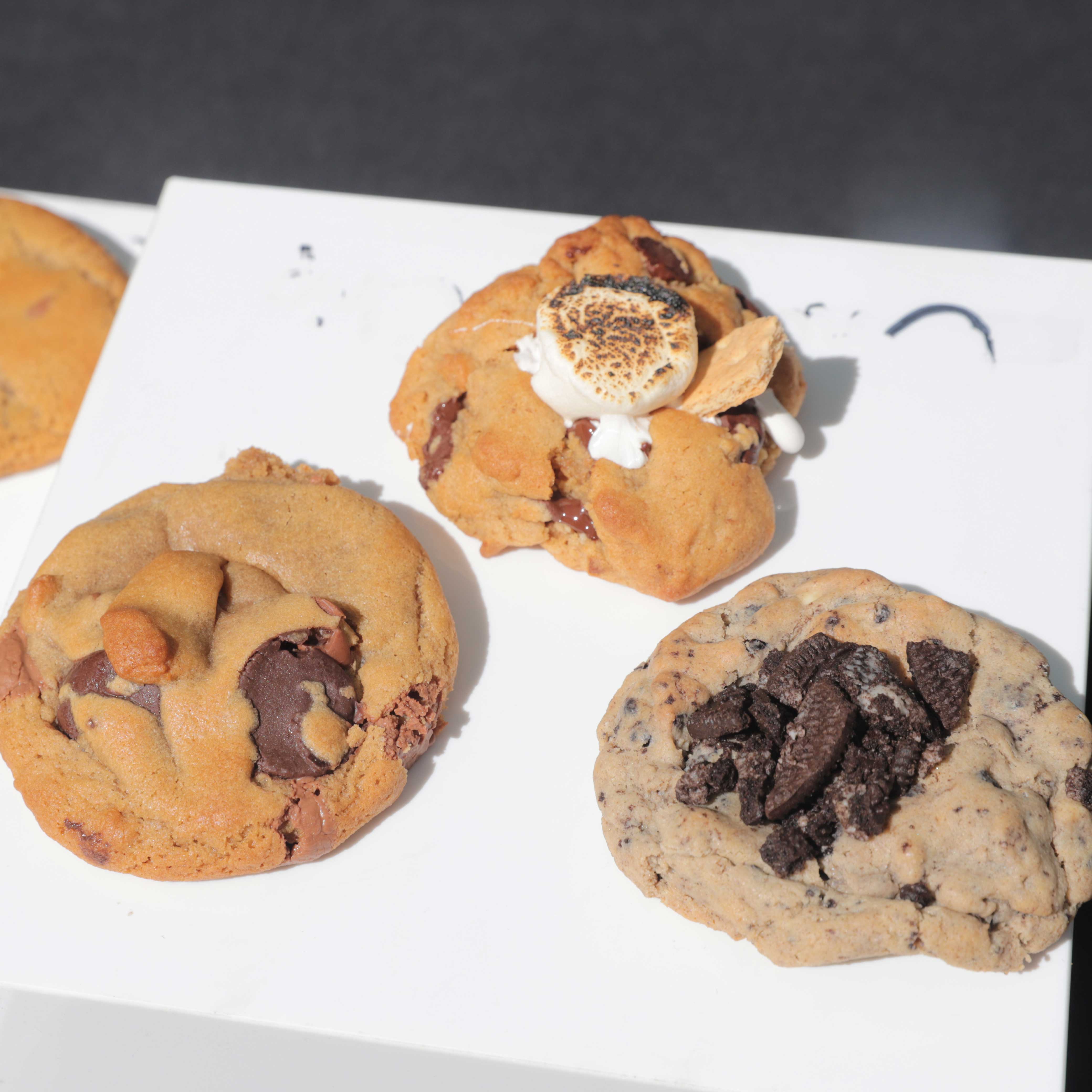 Three large cookies on a white platform. Their toppings depict their flavors: chocolate chip, s'mores, and oreo.