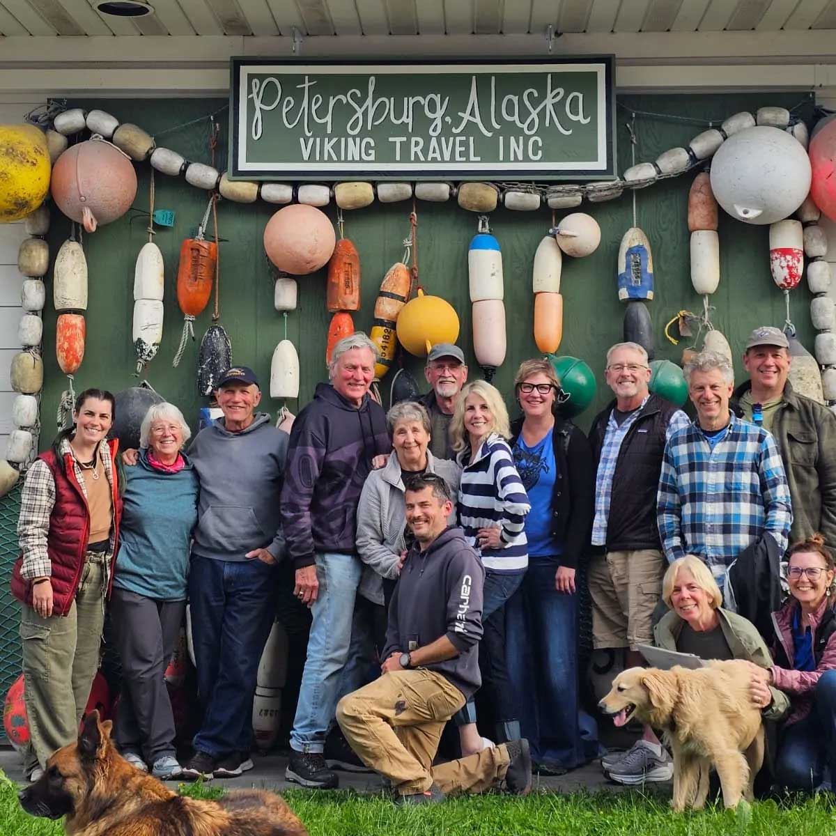 Mark stands with a large group of fellow travelers and dogs under a sign that says 'Petersburg, Alaska.' 