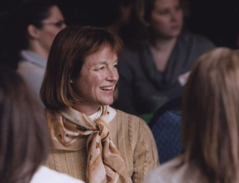 A woman wearing a beige scarf and beige sweater over a white shirt smiles while engaging with a crowd of people.