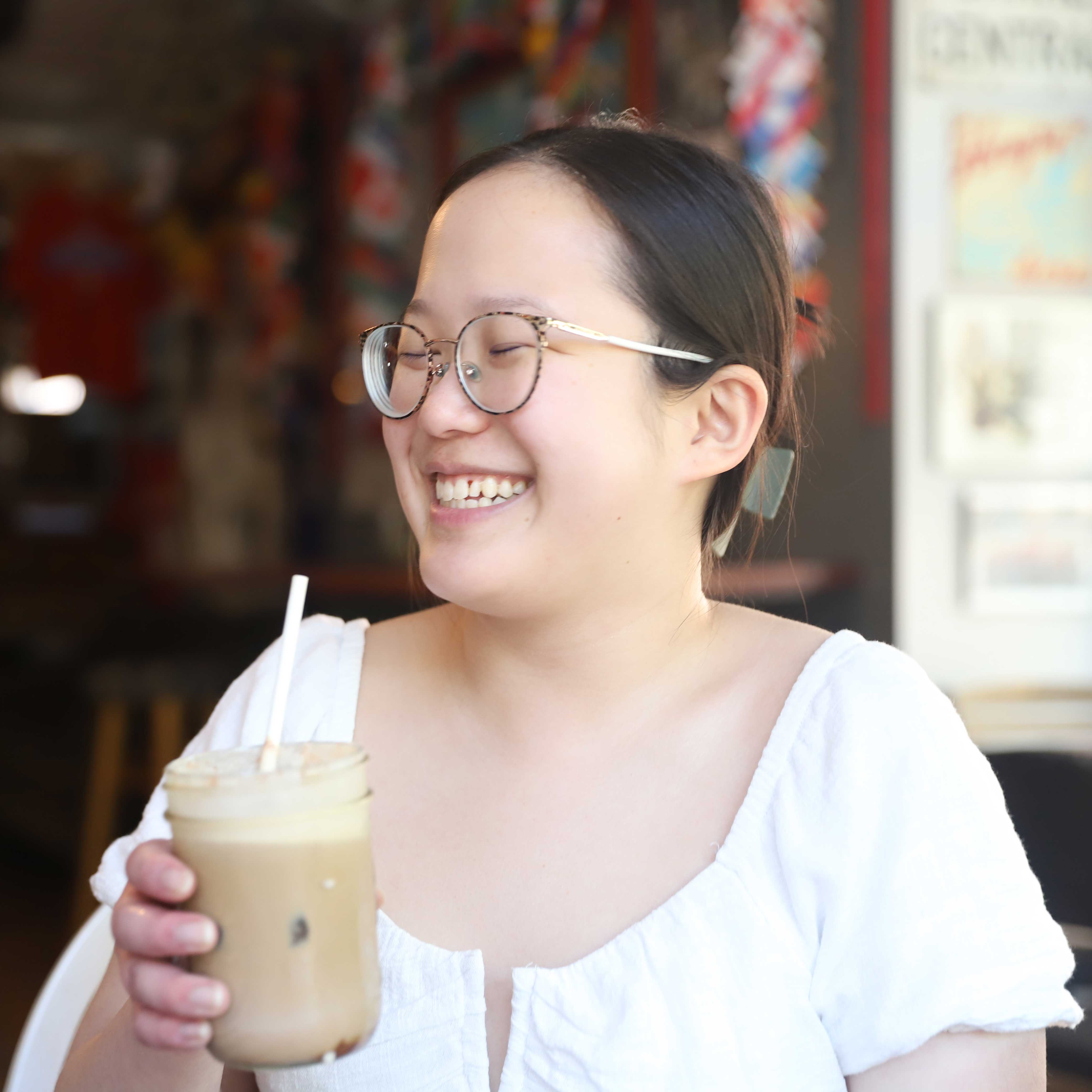 A young woman smiles while holding an iced coffee in her hand. She wears a white top, round glasses, and a claw clip in her hair.