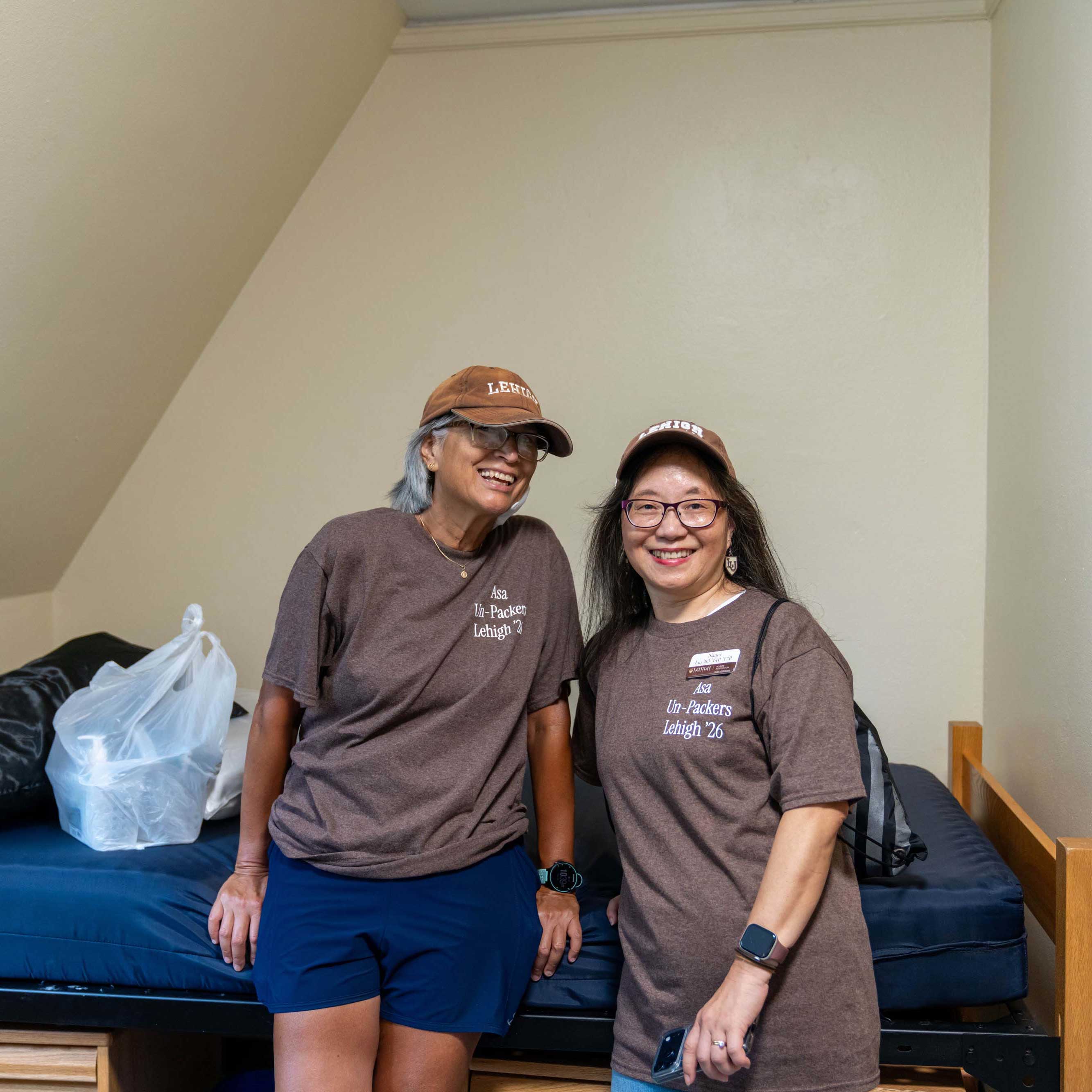 Sandra and Nancy stand inside a dorm room and smile. They wear their brown Asa Un-Packer shirts and Lehigh hats.