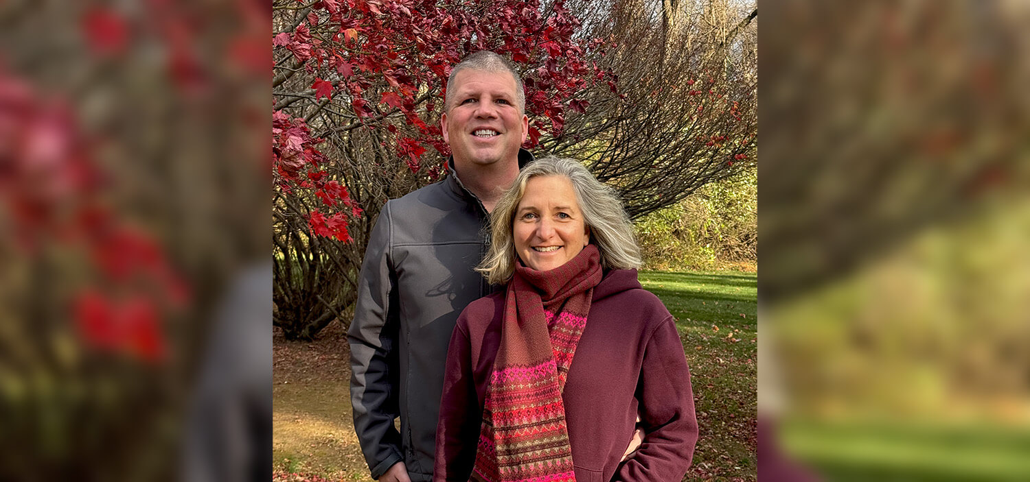 A couple stand closely, smiling, on a chilly and sunny day.