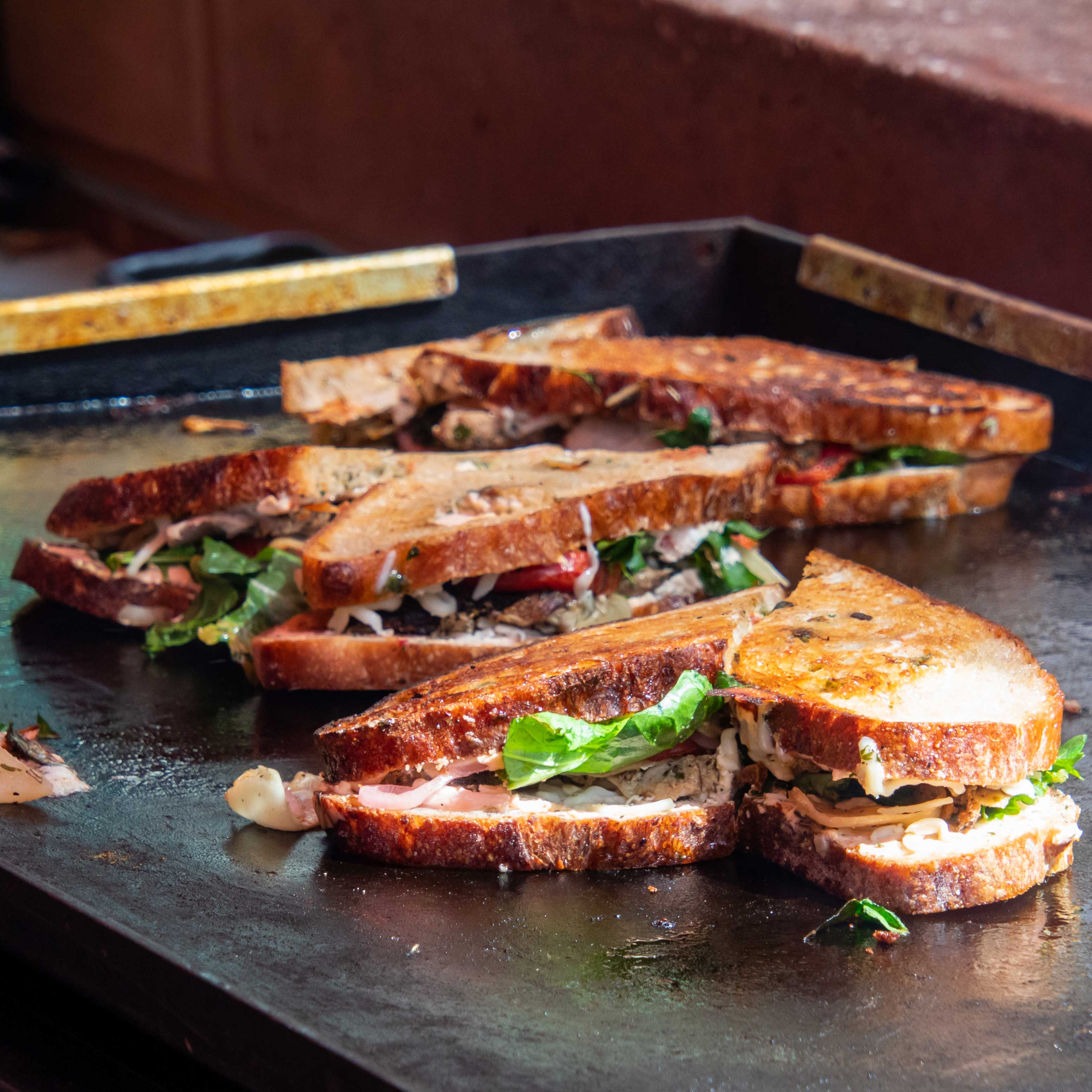 Several sandwiches on a flat-top griddle. The bread is a warm, toasted brown, and greenery and cheese peek out the sides.
