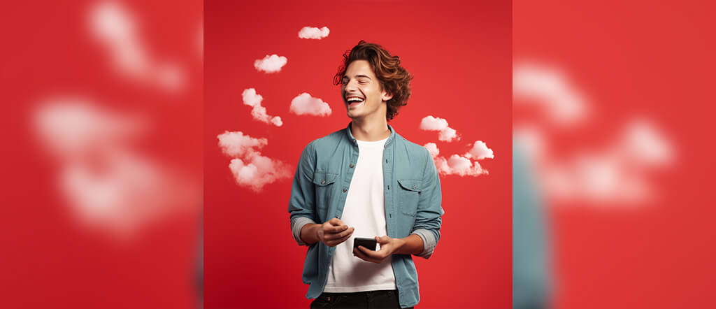 A laughing man is holding a cell phone while standing against a red backdrop wears a white tshirt under a pale blue button down.