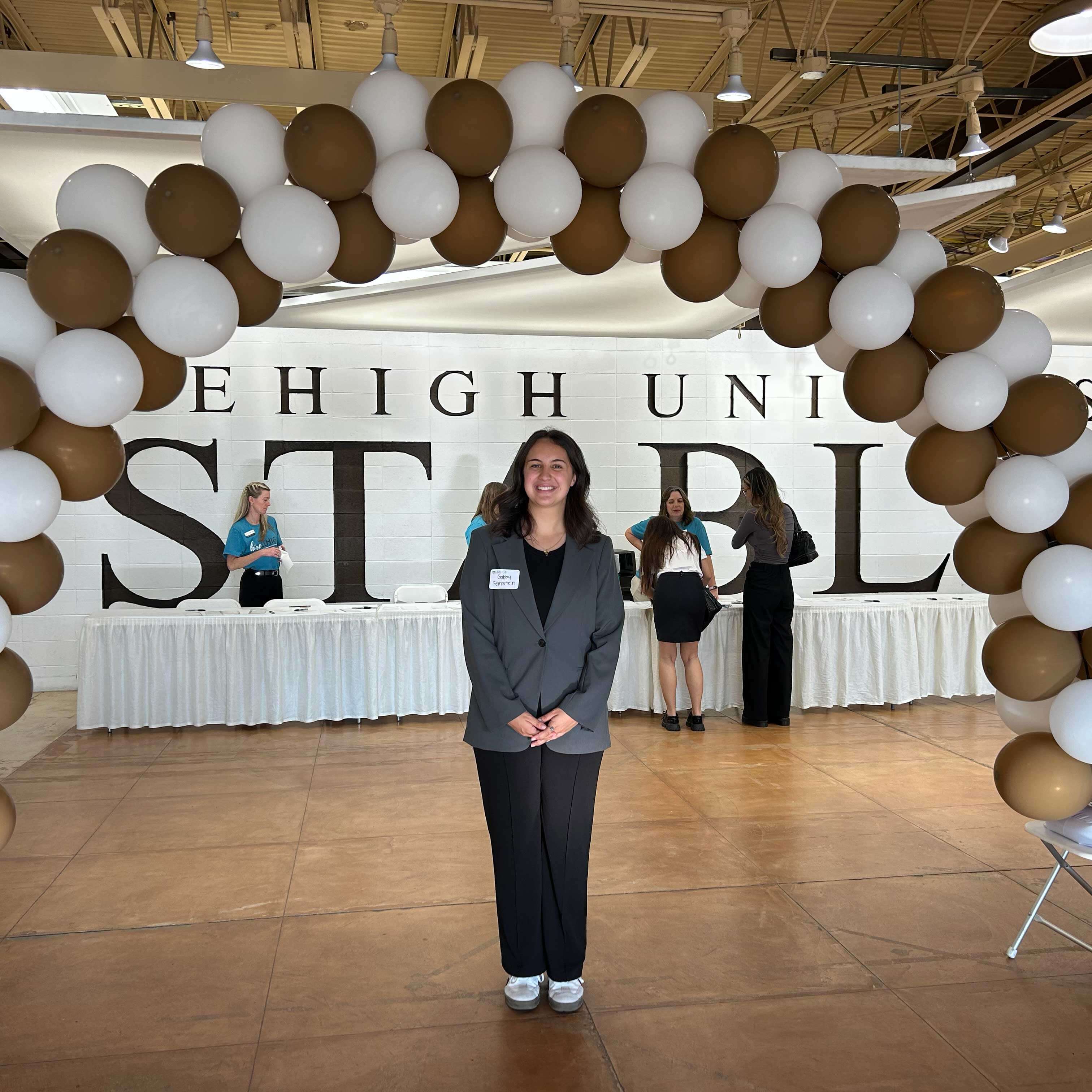 Gabby attends the Career Fair in a dark grey blazer, black trousers, and white sneakers. She smiles and wears a name tag.