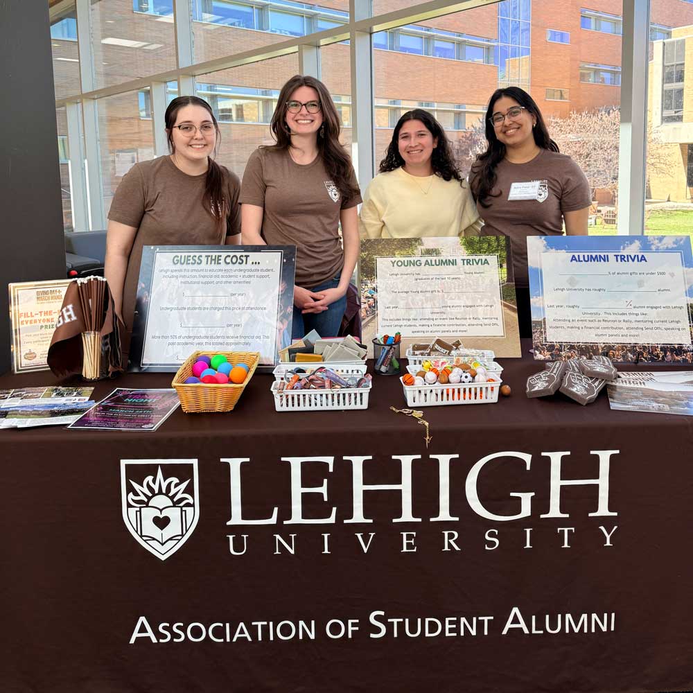 Four women stand smiling behind a table draped with a dark tablecloth featuring the Lehigh University and Association of Student Alumni logos. The table is lined with trivia signs, informational fliers, small giveaway prizes, and baskets of colorful stress balls in front of a glass building wall.