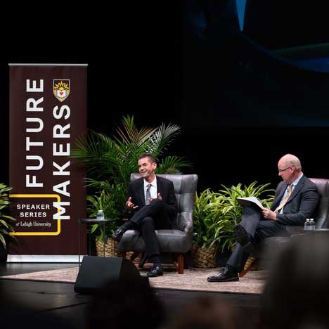 Jared Isaacman wears a black suit and is mid-conversation. He sits beside a Lehigh Future Makers banner.
