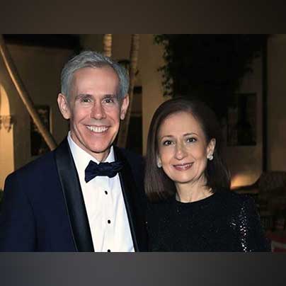 John wears a classic tuxedo and stands with Maria, who wears a black, dress.