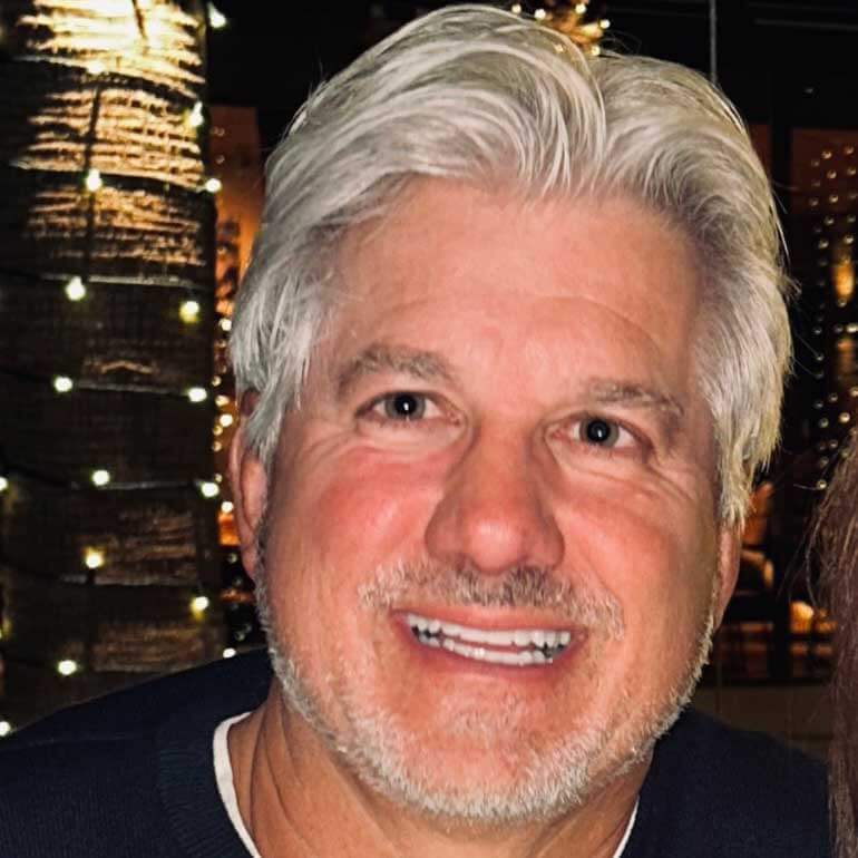 John wears a dark, navy sweater and smiles. Behind him are string lights that paint the photo in a warm, golden glow.