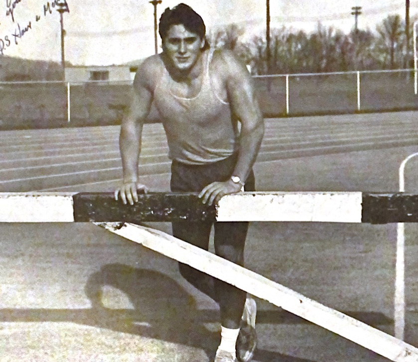 A young muscular man poses on a track field against a hurdle.
