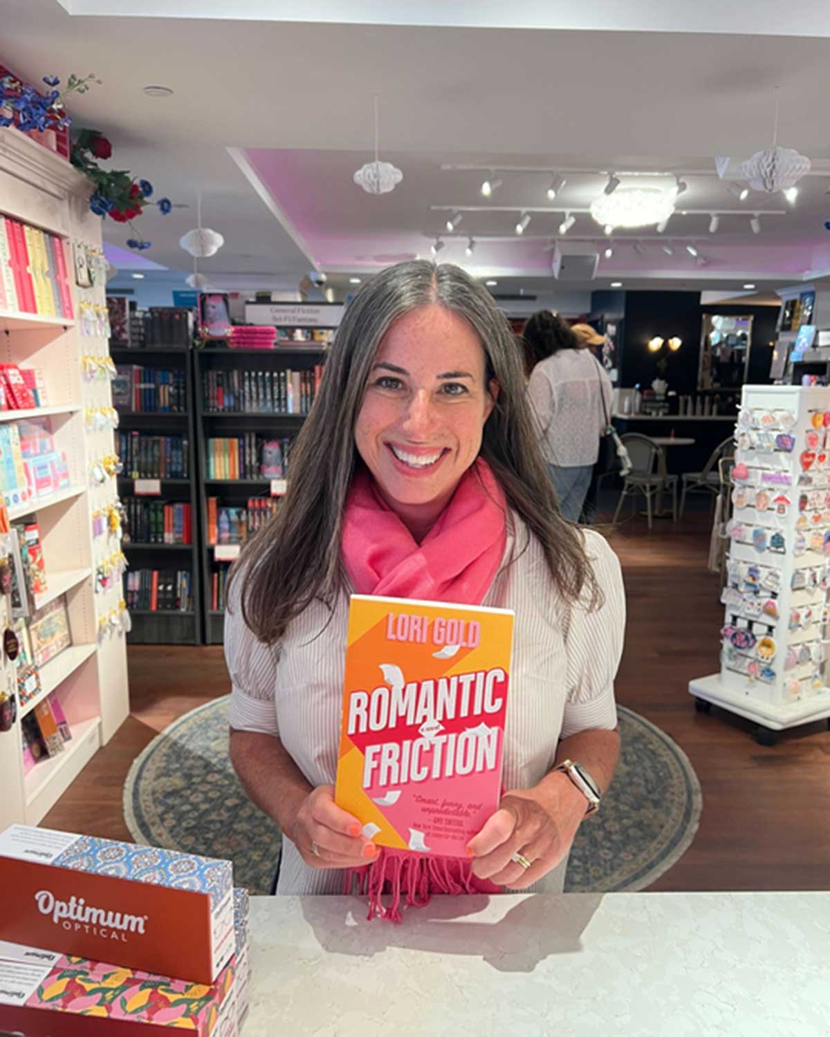 Lori wears a bright pink scarf over a white shirt and holds up Romantic Friction. Behind her, patrons scan bookshelves.