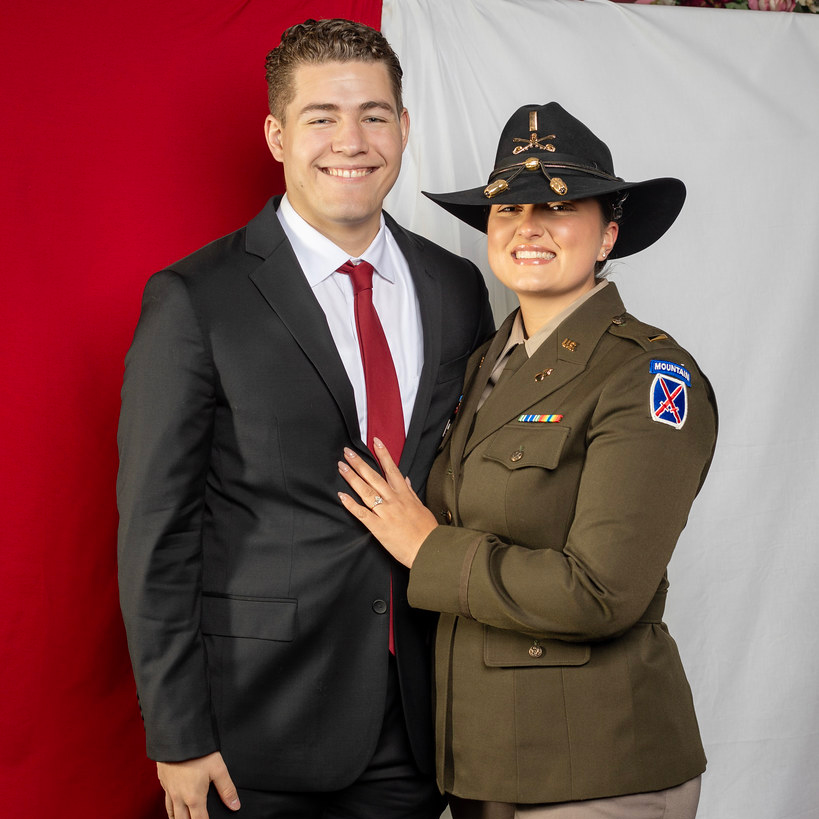 Maura and Ben pose in front of a red and white background. She wears a dark green Ordnance Officer jacket and black hat.