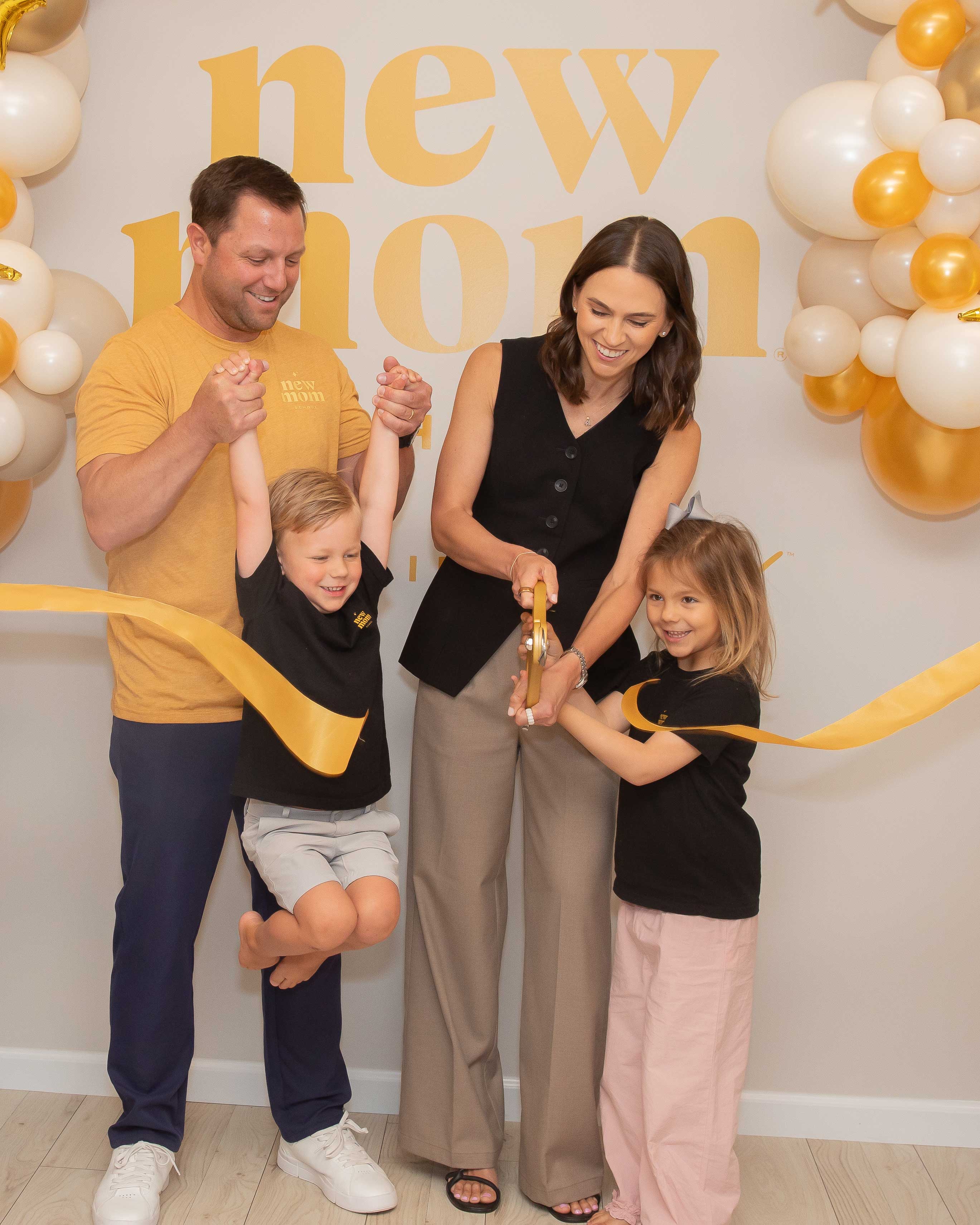 Danielle with her husband and children cutting a yellow ribbon for the opening of New Mom School. They smile and wear merch.