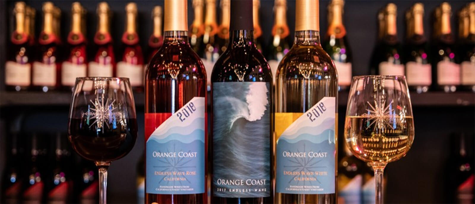 Three bottles of orange coast wine in between two wine glasses atop a wooden countertop.