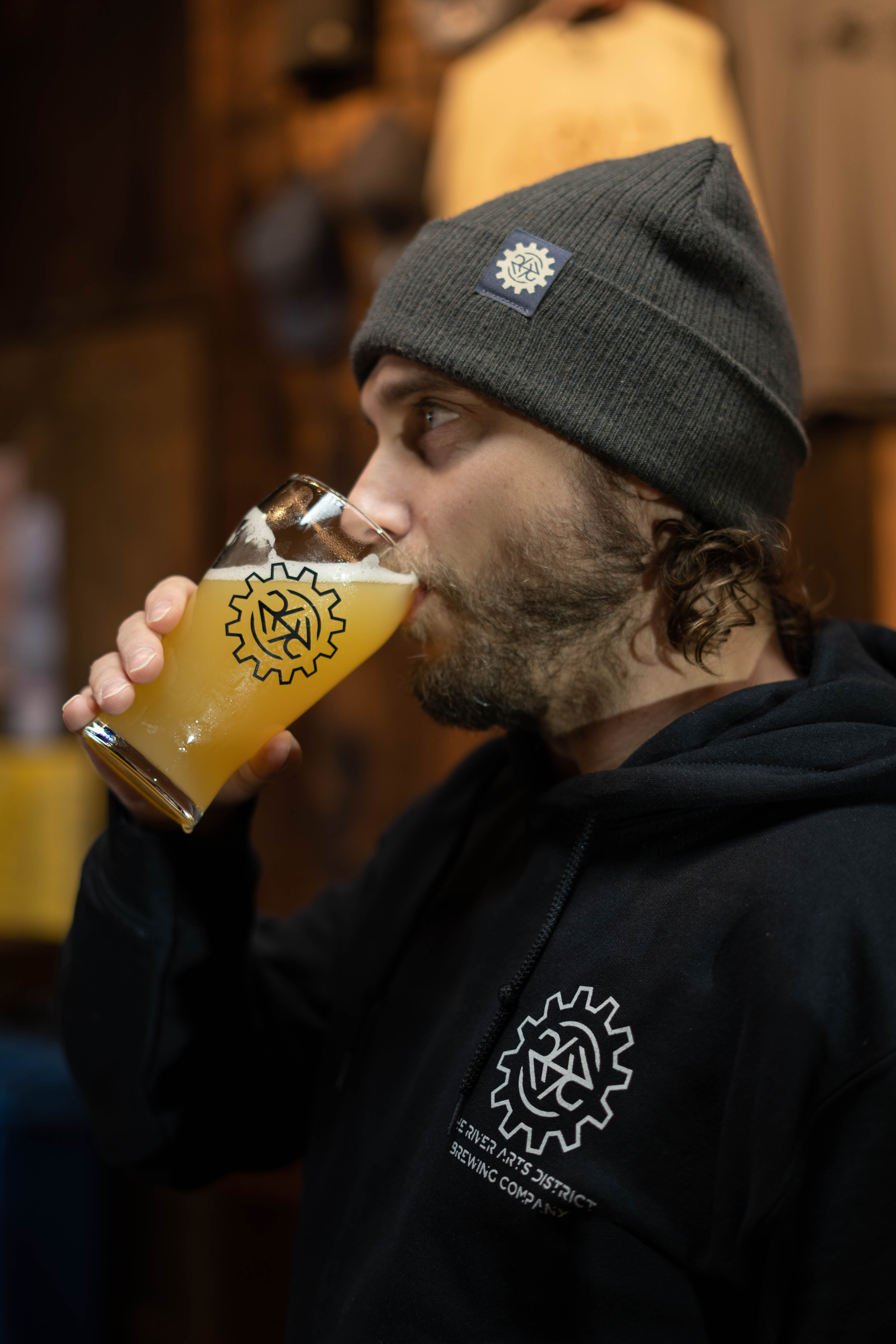 Robert Lemery drinks a beer with his brewery's logo on the glass, his sweatshirt, and hat.