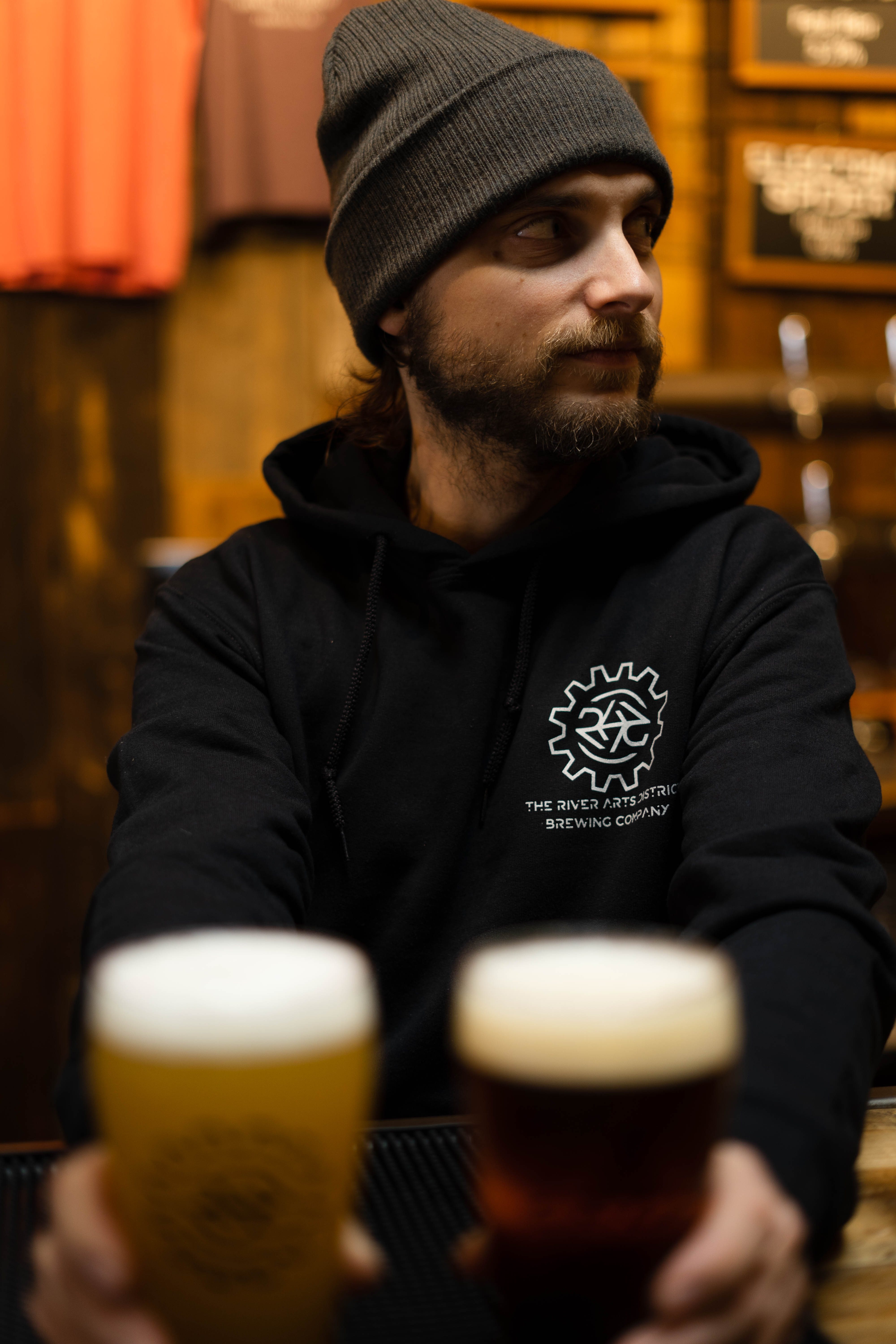 Lemery holds two glasses of beer of different colors wearing a  black sweatshirt with his brewery's logo on it